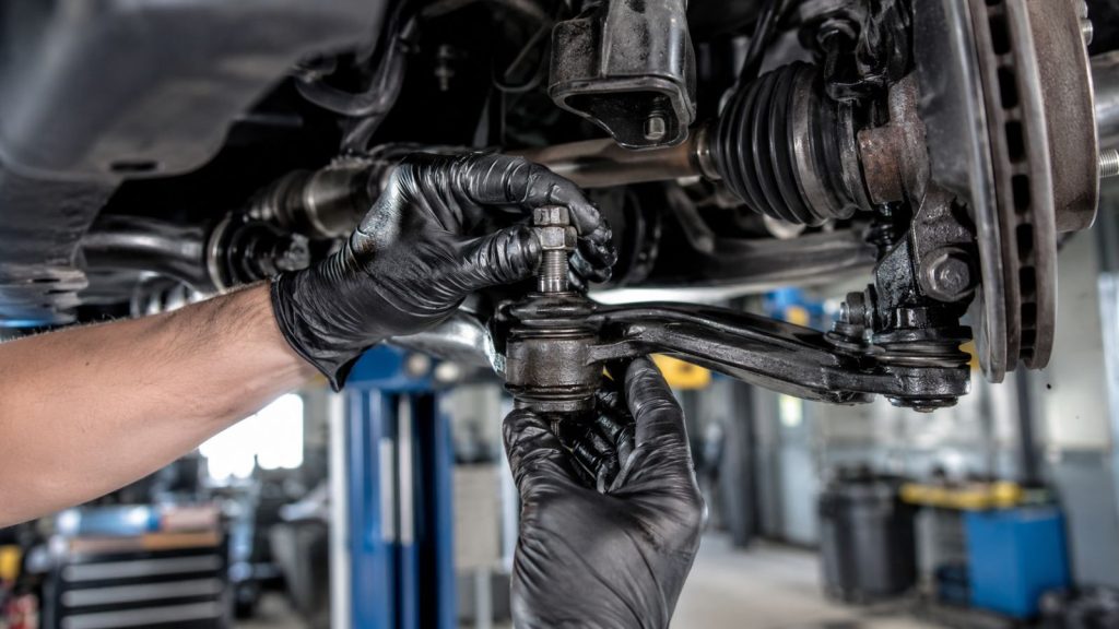 A professional mechanic in an auto repair shop performing a hands-on diagnostic inspection of a vehicle's front suspension ball joint and tie rod assembly.