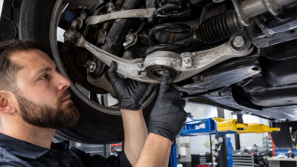 A technician at an auto repair shop performing a suspension bushing play inspection on a vehicle raised on a car lift.