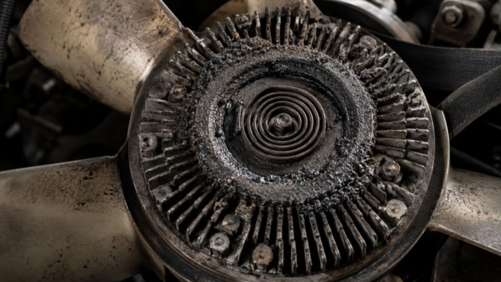 A close-up view of a failed engine viscous fan clutch assembly, showing a dark, circular ring of greasy road grime and dust coating the bimetallic spring. This residue is the visual indicator of a blown silicone fluid seal that caused overheating during low-velocity idling.
