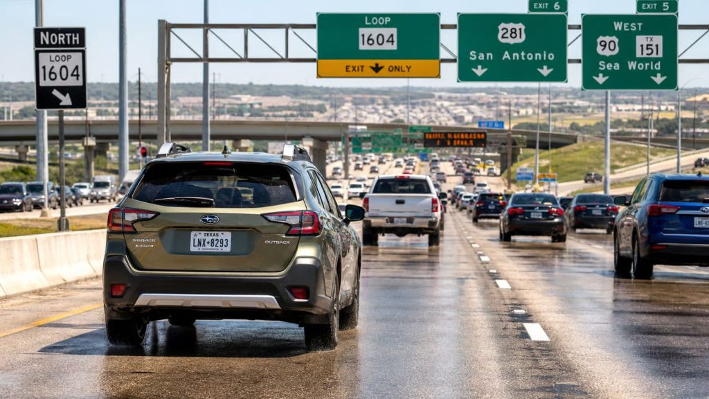 A silver Subaru navigating heavy stop-and-go traffic on the Loop 1604 interchange in Alamo Ranch, Texas, under the intense summer sun with visible heat shimmer rising from the highway.
