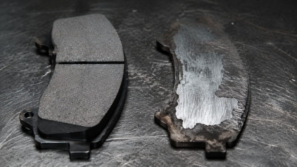 Side-by-side macro comparison of a healthy, thick brake pad versus a severely worn, uneven brake pad on a professional automotive workbench.